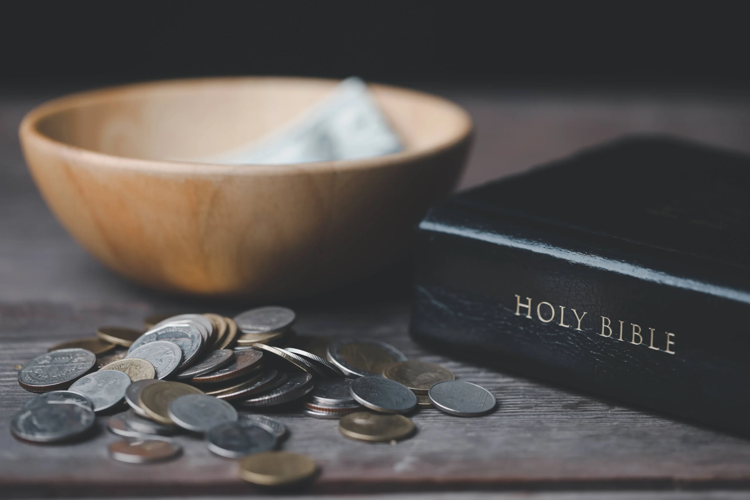 A wooden bowl with paper money, coins scattered on a table, and a closed Bible nearby.