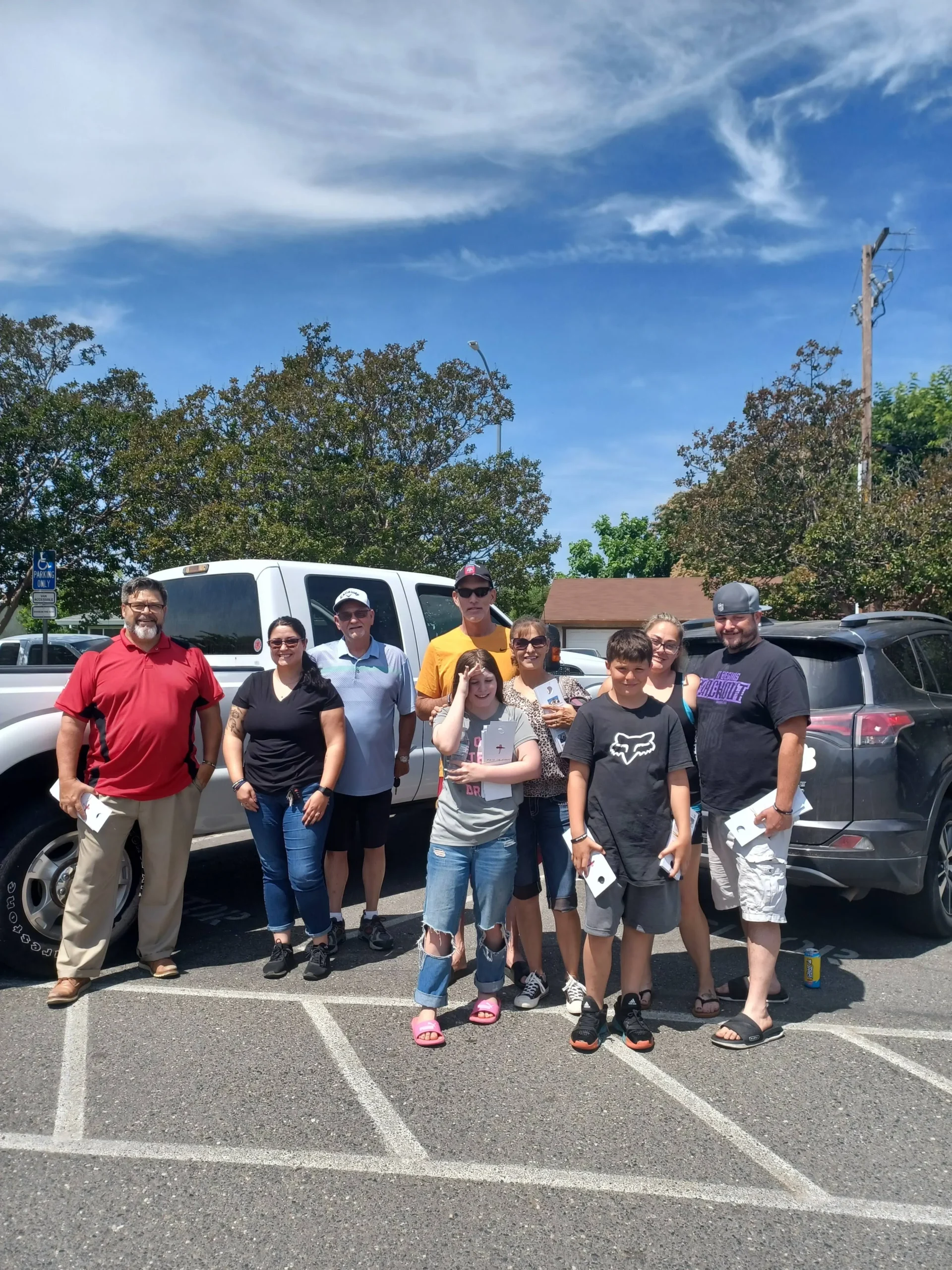 A group of people standing in a parking lot with cars and trees in the background, under a blue sky with some clouds.