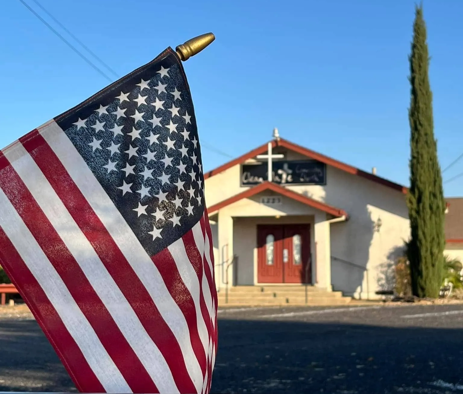 An American flag in the foreground with a church and a tall cypress tree in the background under a clear blue sky.