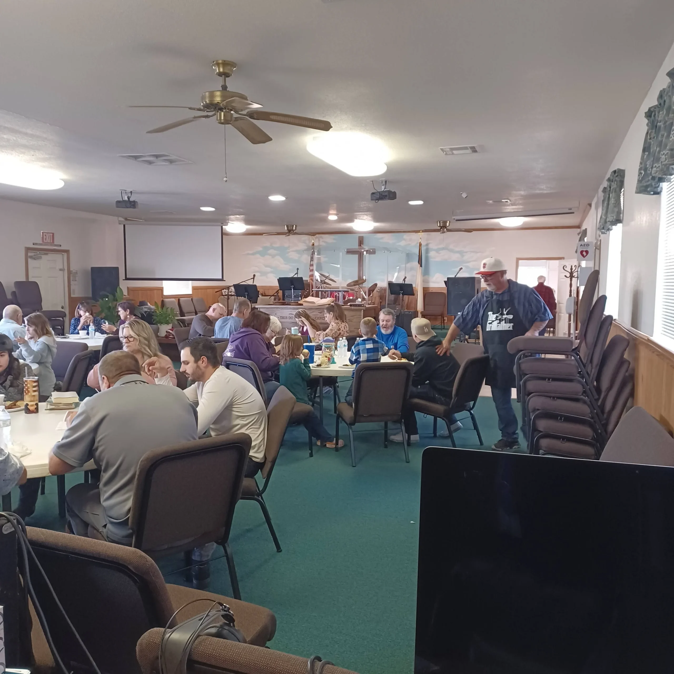 People seated at tables in a community hall, engaging in conversation and activities. The room has a mural, rectangular tables, chairs, and ceiling fans.