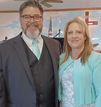 A man and woman stand side by side in a church, both smiling. The man wears a suit with a turquoise tie, and the woman wears a turquoise cardigan over a floral dress.