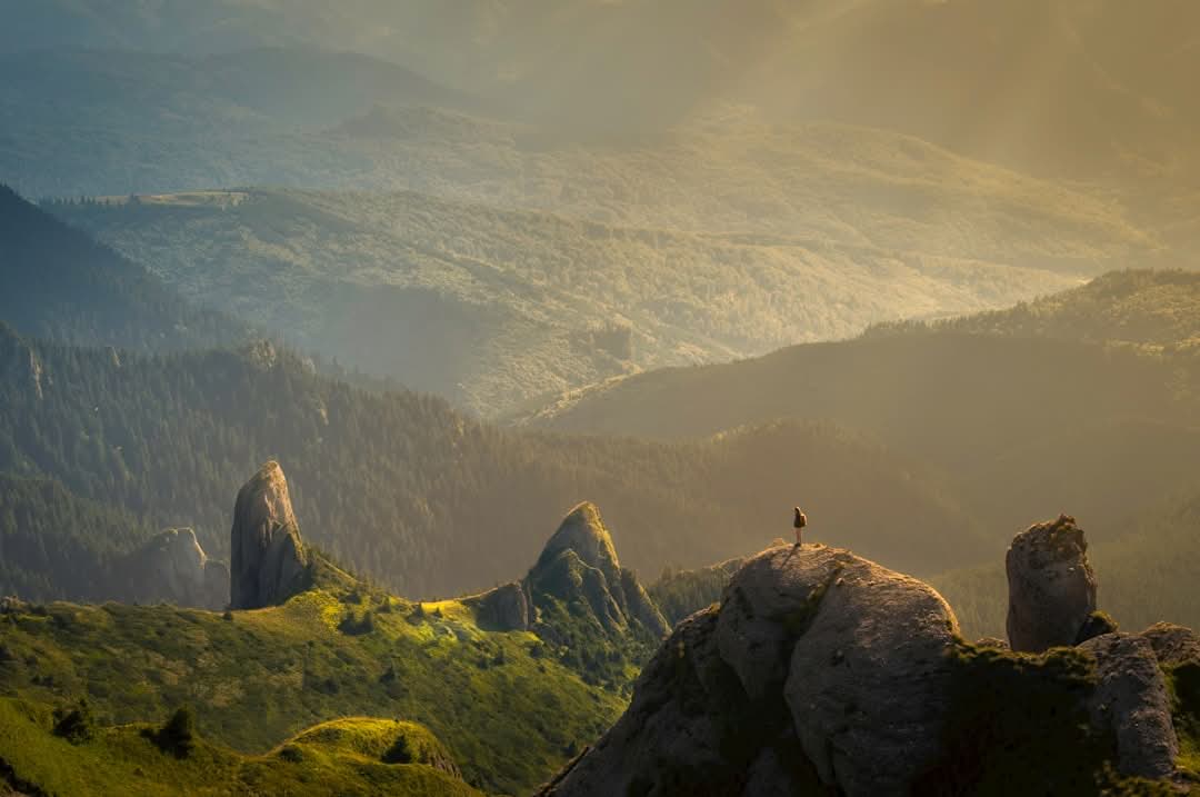 A person stands on a rocky peak overlooking rolling green hills and distant forested mountains under soft, golden sunlight.