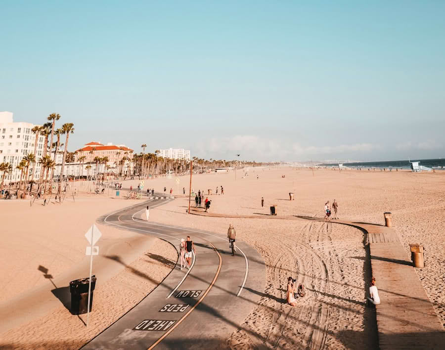 Wide sandy beach with people walking, biking, and sitting; paved path curves through the scene; palm trees and buildings in the background under a clear blue sky.