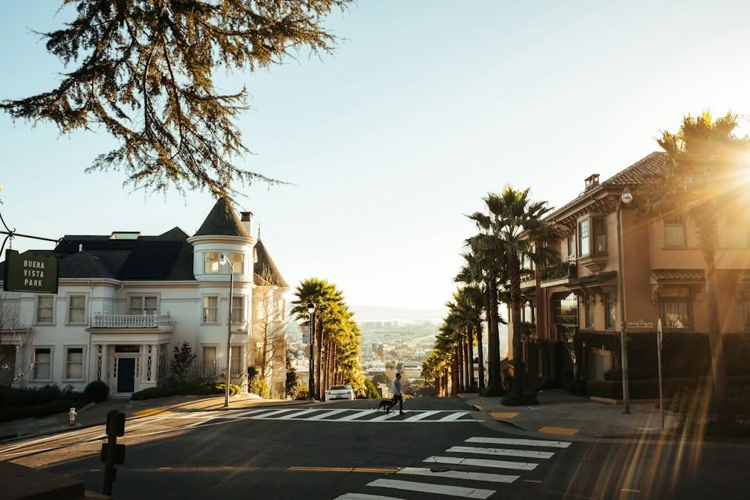 City street with palm trees, a white Victorian-style house, and a person crossing at a crosswalk in sunlight.