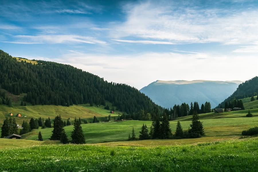 Green grassy valley with scattered trees and small houses, surrounded by forested hills and distant mountains under a partly cloudy blue sky.