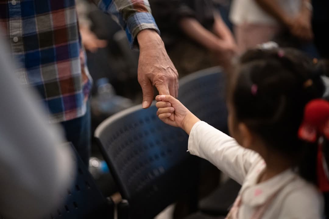 An adult and a child hold hands while standing among rows of black chairs. An adult and a child hold hands while standing among rows of black chairs.